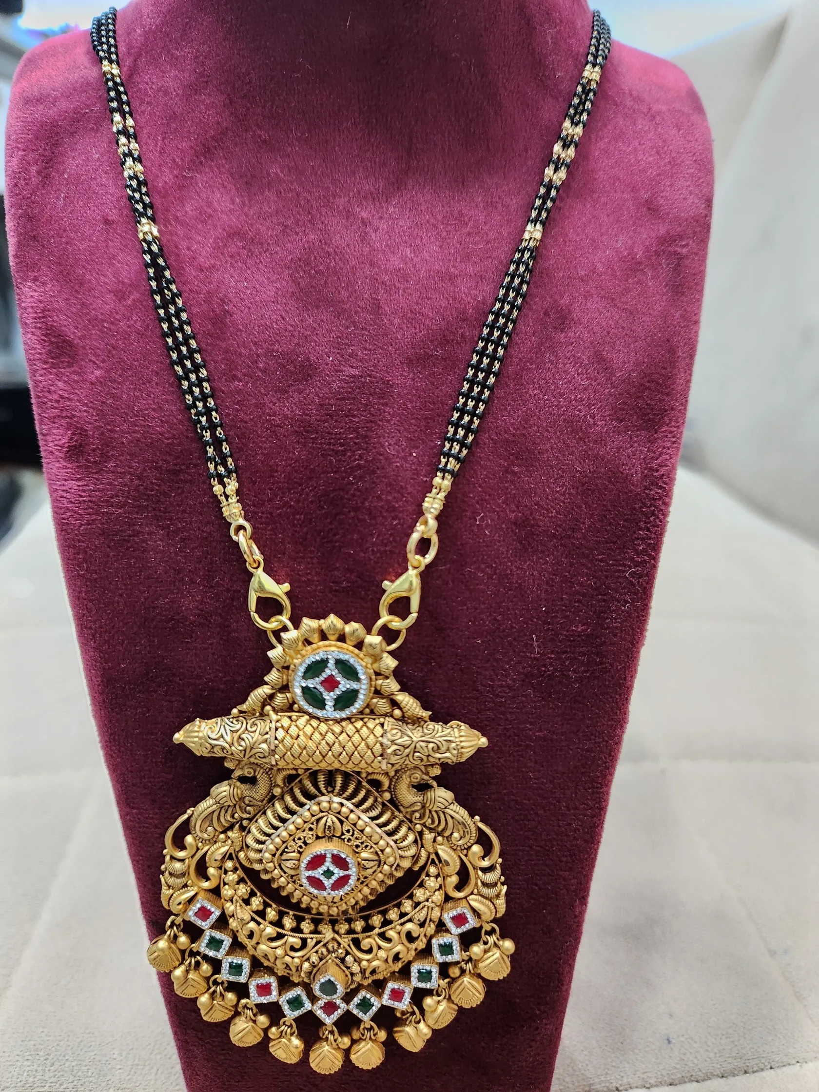 Mangalsutra with double black-bead chain and gold pendant of twin peacocks, red and green stones, on a maroon bust