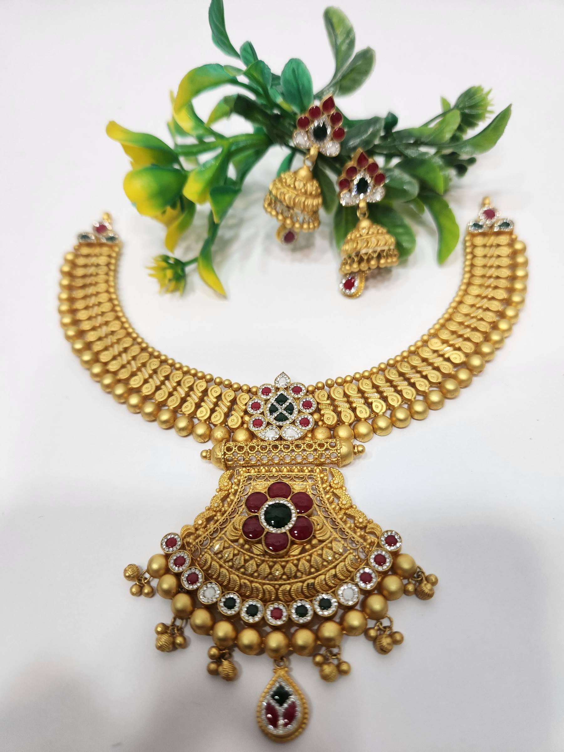 Matte gold collar necklace with fan-shaped pendant, red and green stones, bell fringe and matching jhumkas